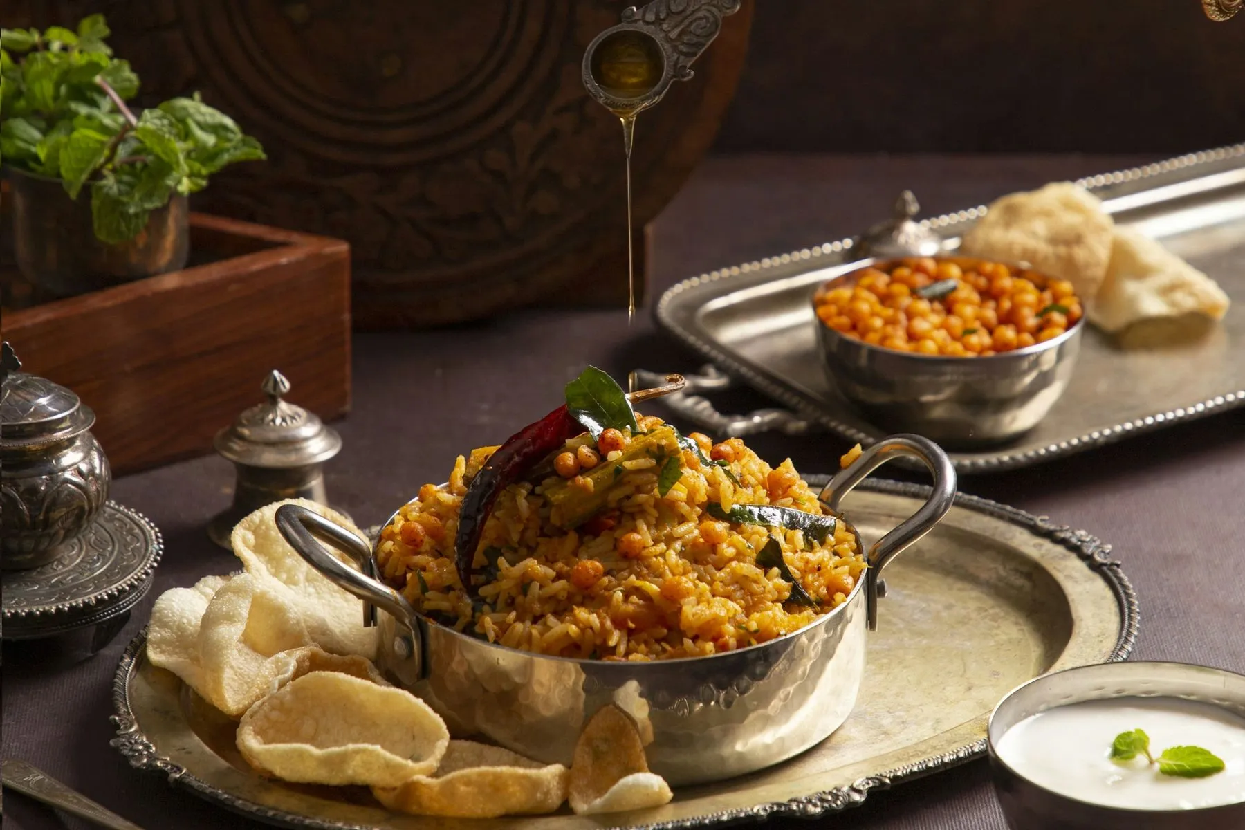 Steel pot of spiced rice-and-lentil dish topped with a dried chili, with ghee being poured from a spoon, papads, boondi and a bowl of yogurt