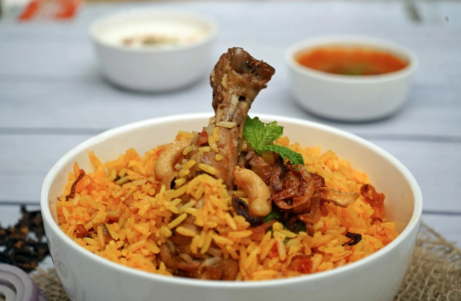 Bowl of saffron-orange biryani rice topped with a chicken leg, with small bowls of raita and curry behind
