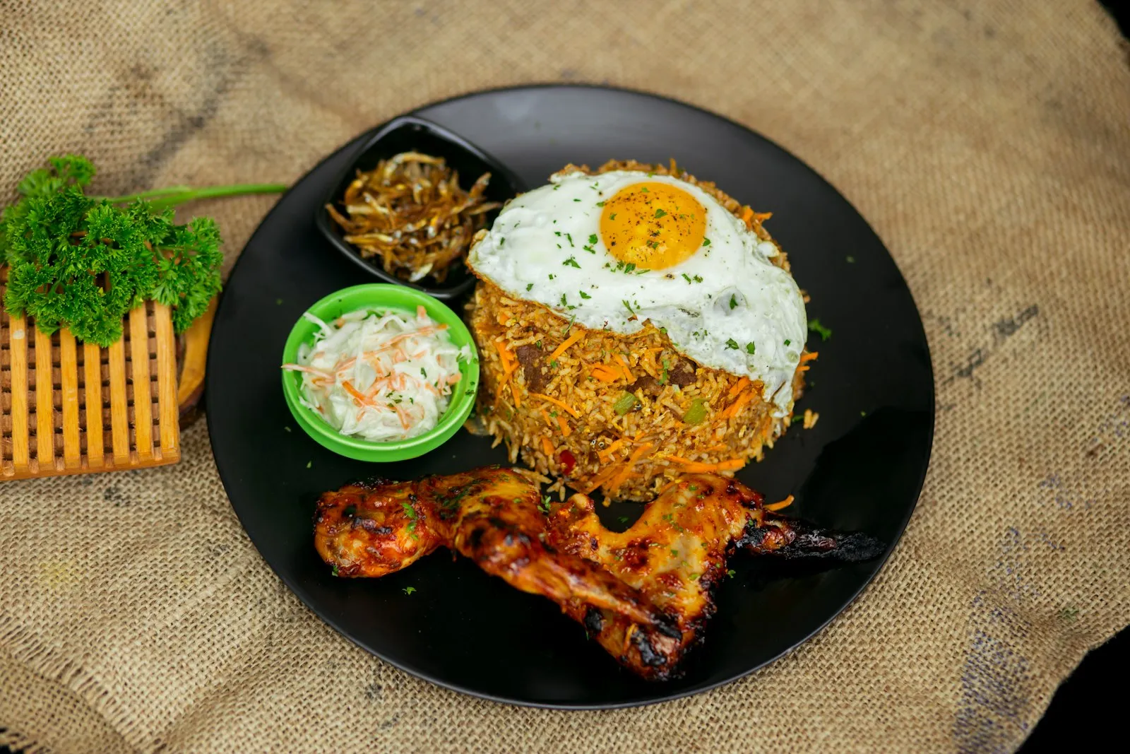 Black plate of fried rice with a fried egg, grilled chicken leg and a small salad