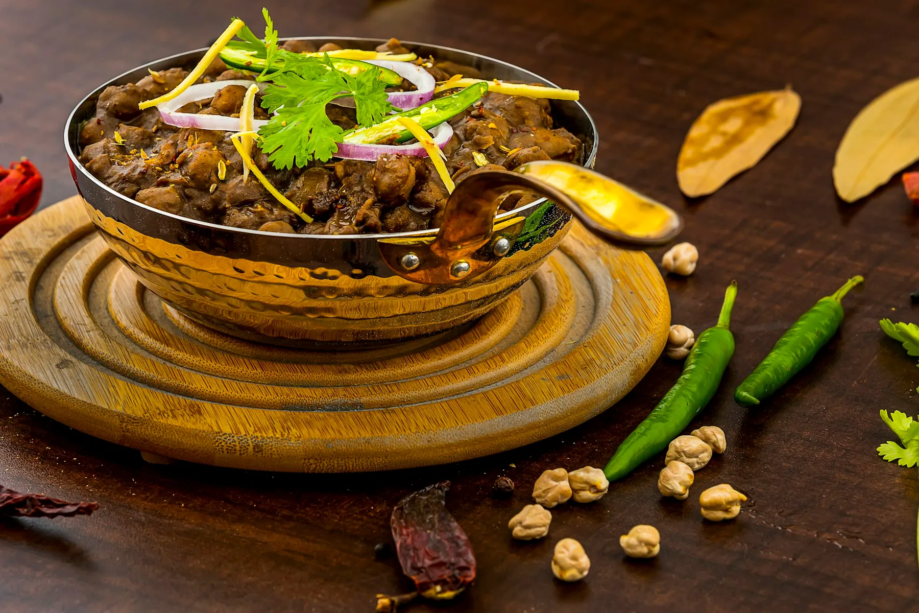 Brass kadai of dark chickpea curry topped with onion rings, ginger and cilantro, with green chilies and chickpeas around it