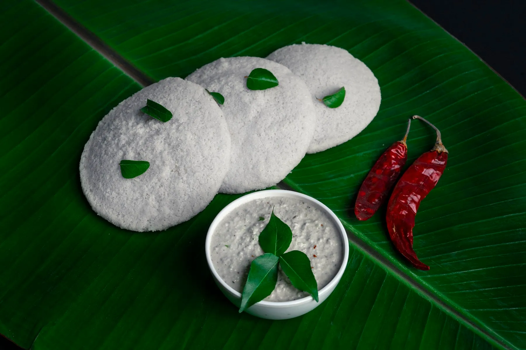 Bowl of white coconut chutney with curry leaves beside three idlis and dried red chilies on a banana leaf