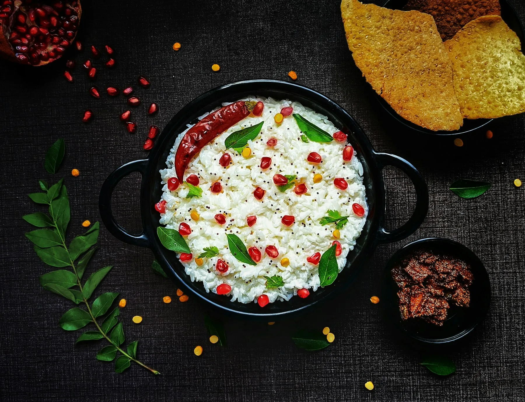 Black pot of curd rice topped with pomegranate seeds, curry leaves and a red chili, with papads and pickle on a dark table