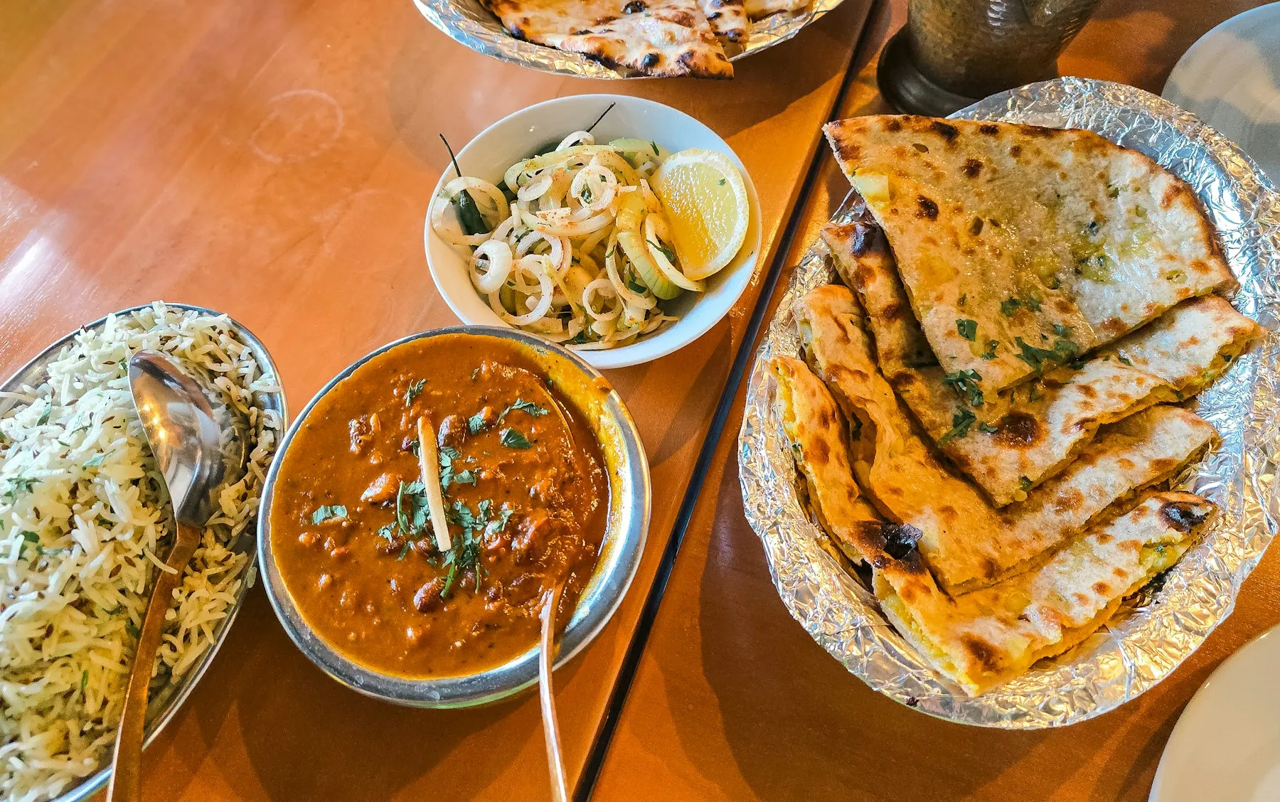 Steel bowl of dark dal makhani with jeera rice, stuffed flatbread and an onion salad on a restaurant table