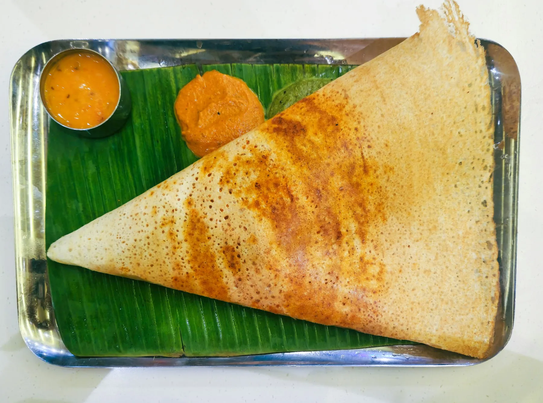 Large triangular crisp dosa on a banana leaf in a steel tray, with sambar and a red chutney