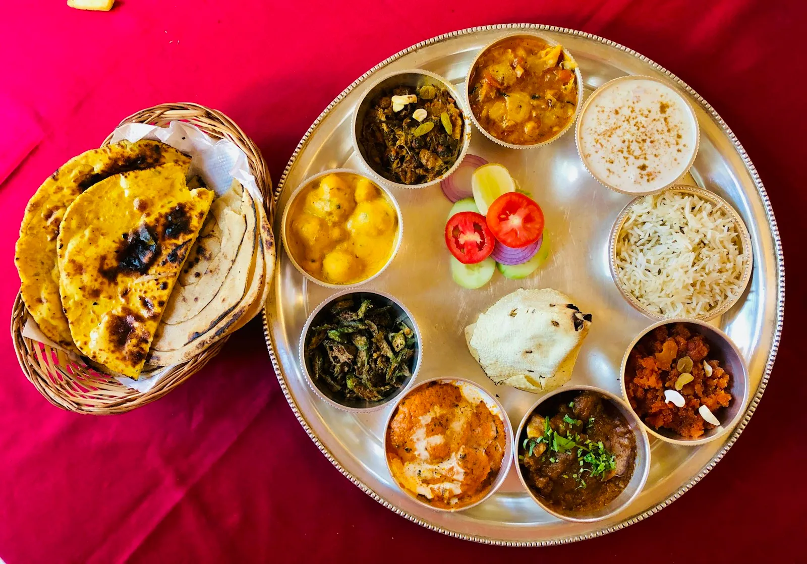 Steel thali with many small bowls of Indian curries, rice and flatbreads on a magenta cloth