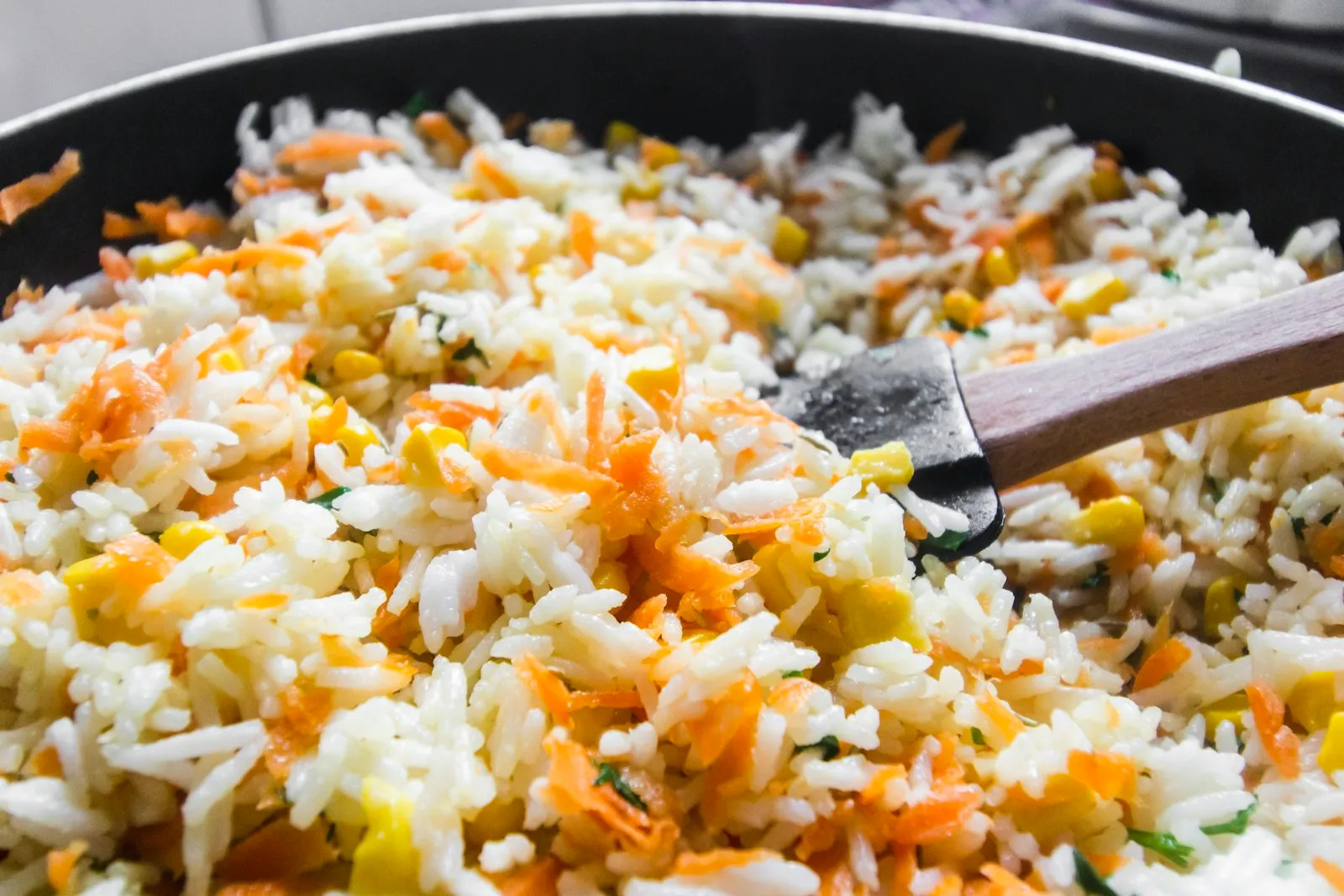 Fried rice with carrots and corn cooking in a black pan with a wooden spatula