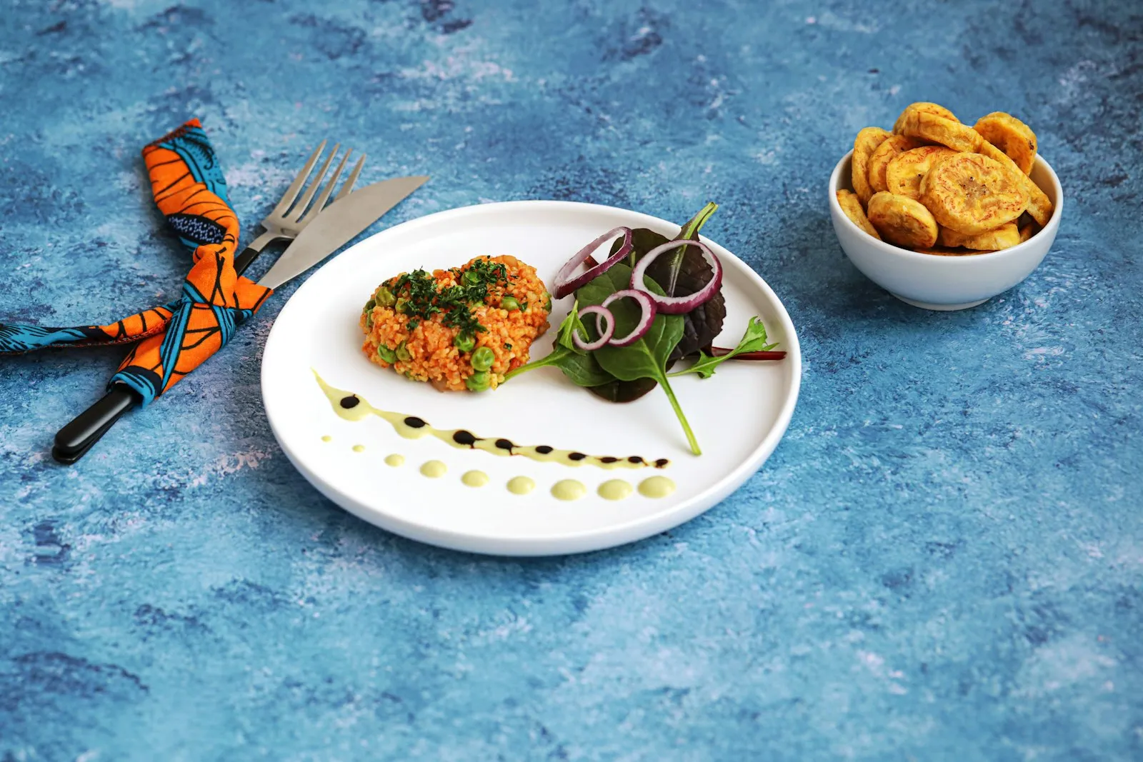 Plate of jollof rice with salad beside a bowl of fried plantain on a blue surface