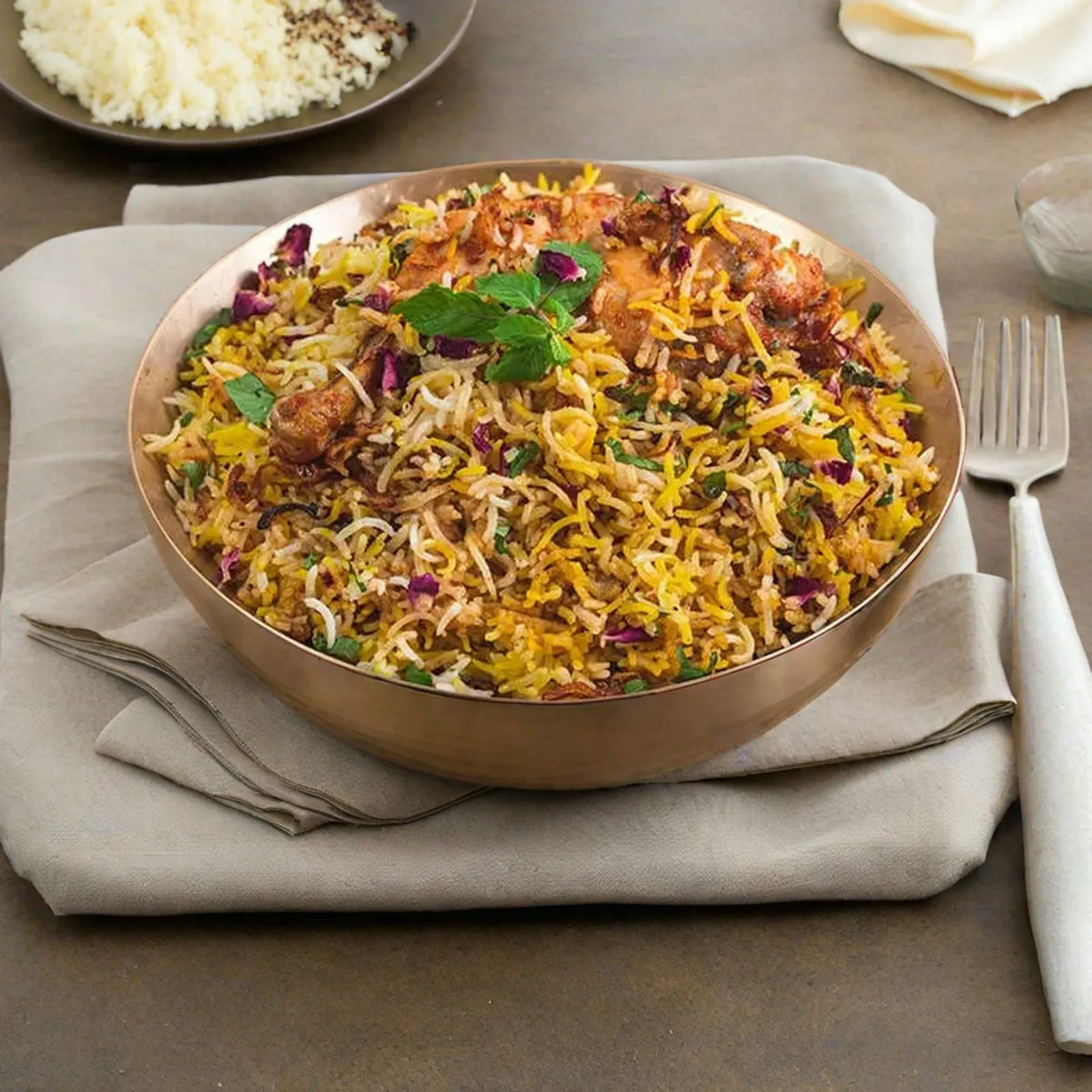 Copper bowl of meat biryani with herbs and fried onions on a linen napkin