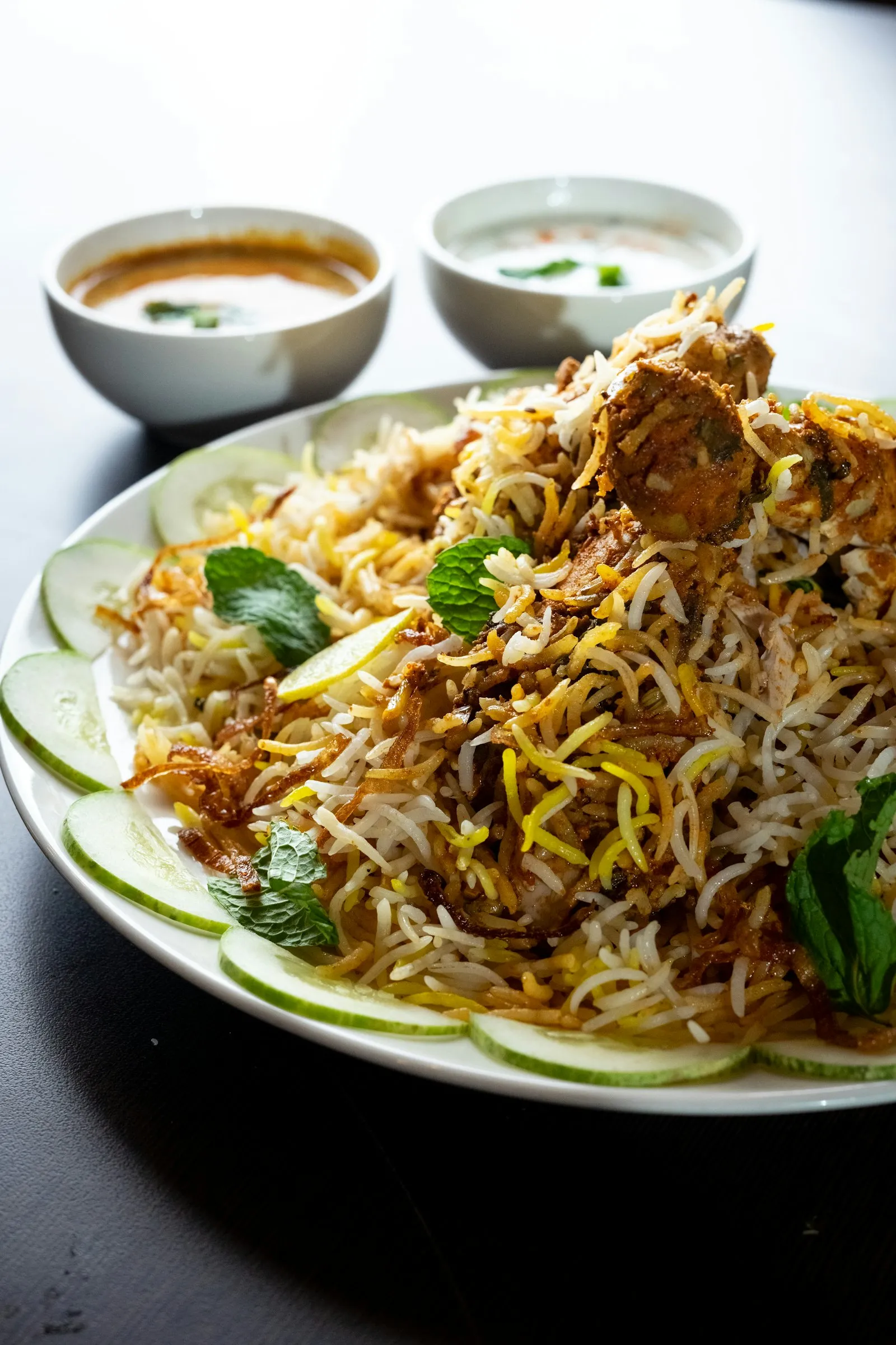 Plate of chicken biryani with long-grain rice, mint and lime slices, with small bowls of raita and gravy