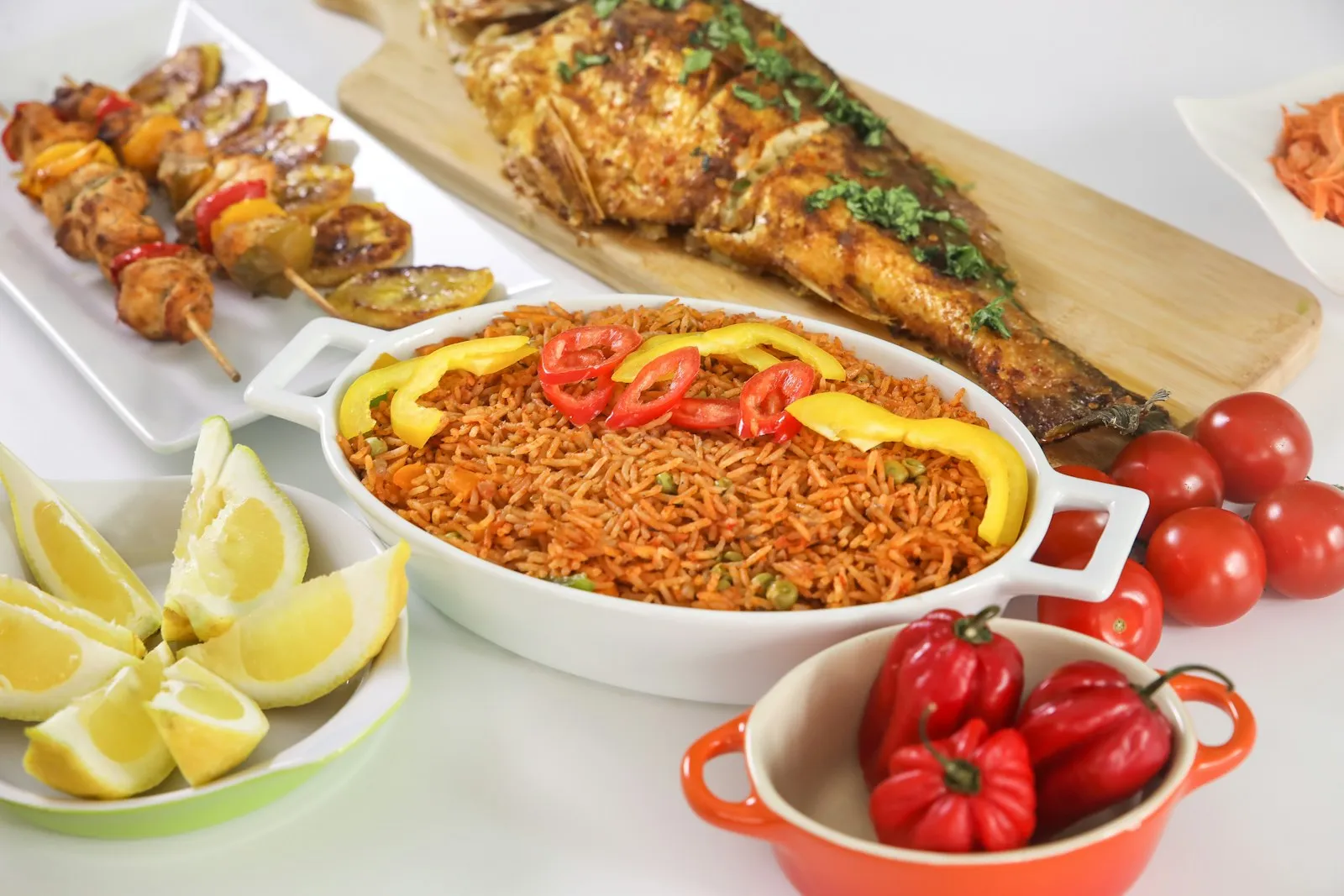 Jollof rice in a white dish with grilled fish, skewers, lemons and scotch bonnet peppers