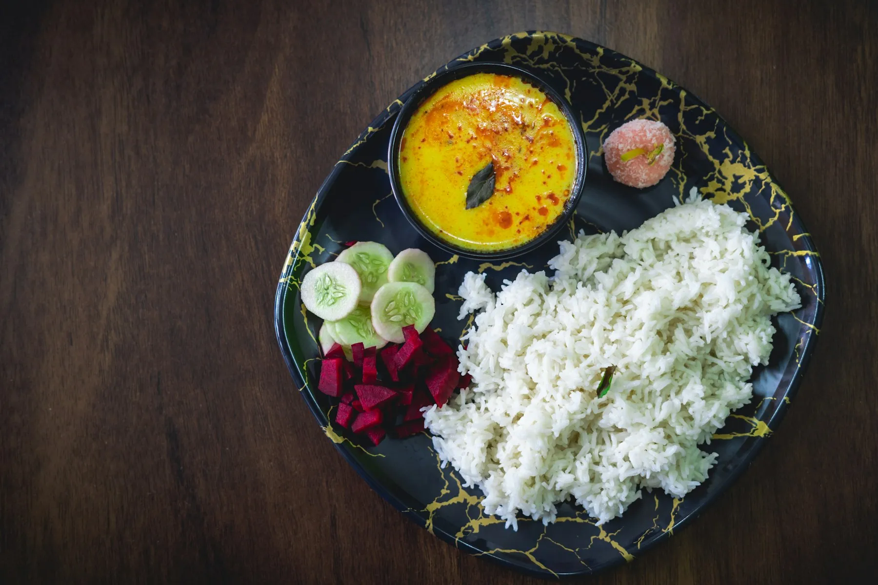 Black plate with a bowl of yellow kadhi, steamed rice, cucumber and beetroot slices and a small sweet
