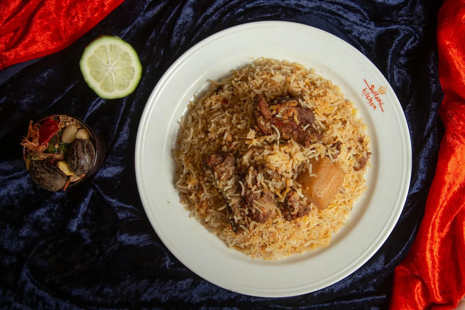 White plate of spiced rice with pieces of meat and potato on a dark cloth