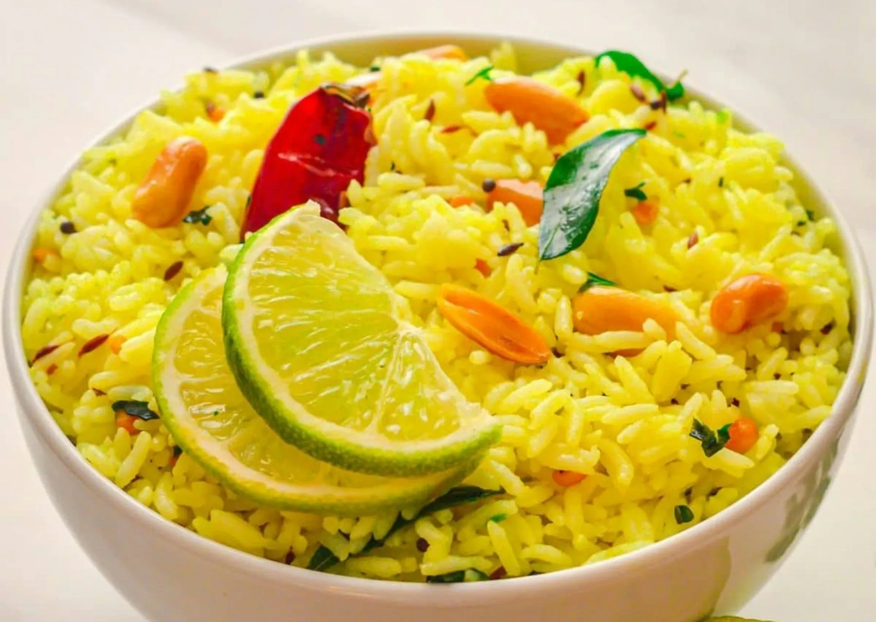 Bowl of bright yellow rice with peanuts, curry leaves, a dried red chili and lime slices
