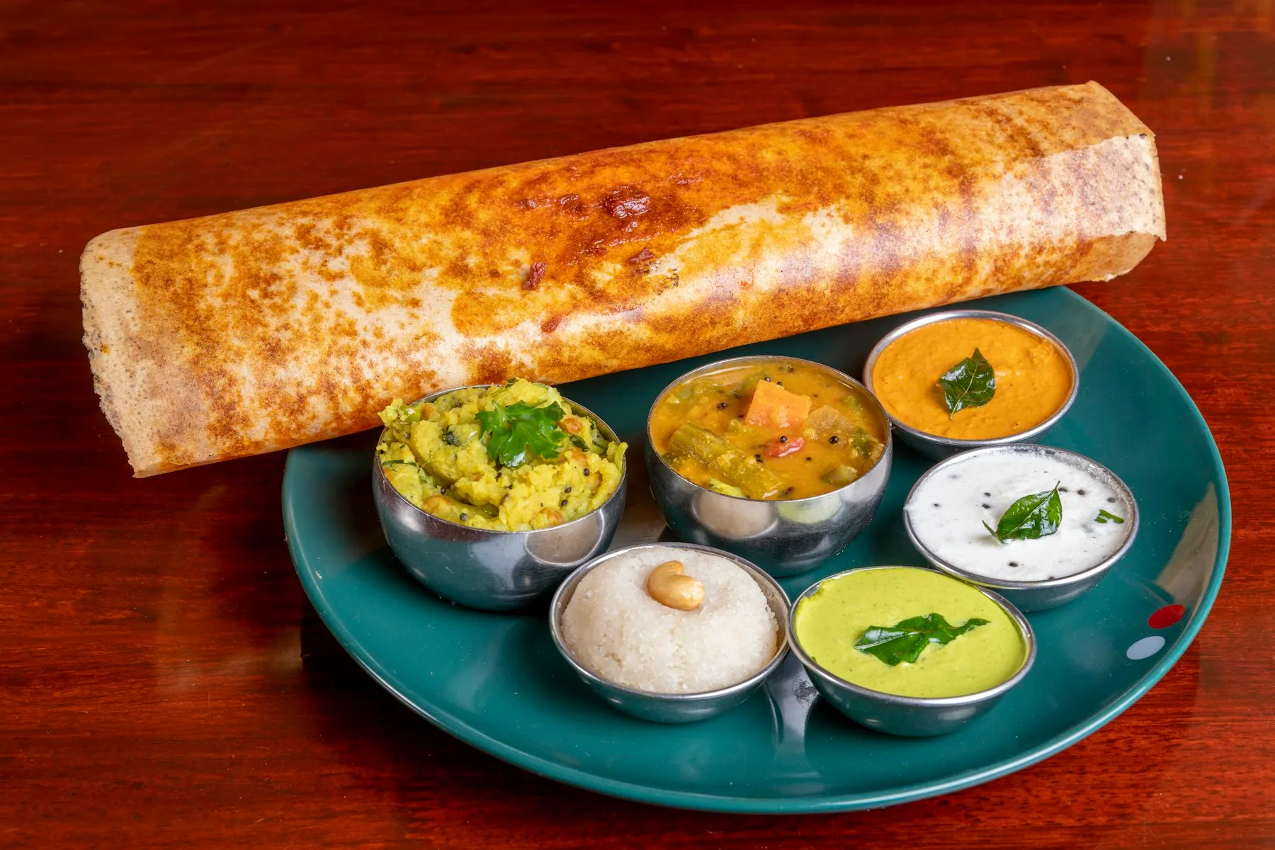 Rolled golden dosa on a teal plate with small steel bowls of potato masala, sambar and chutneys