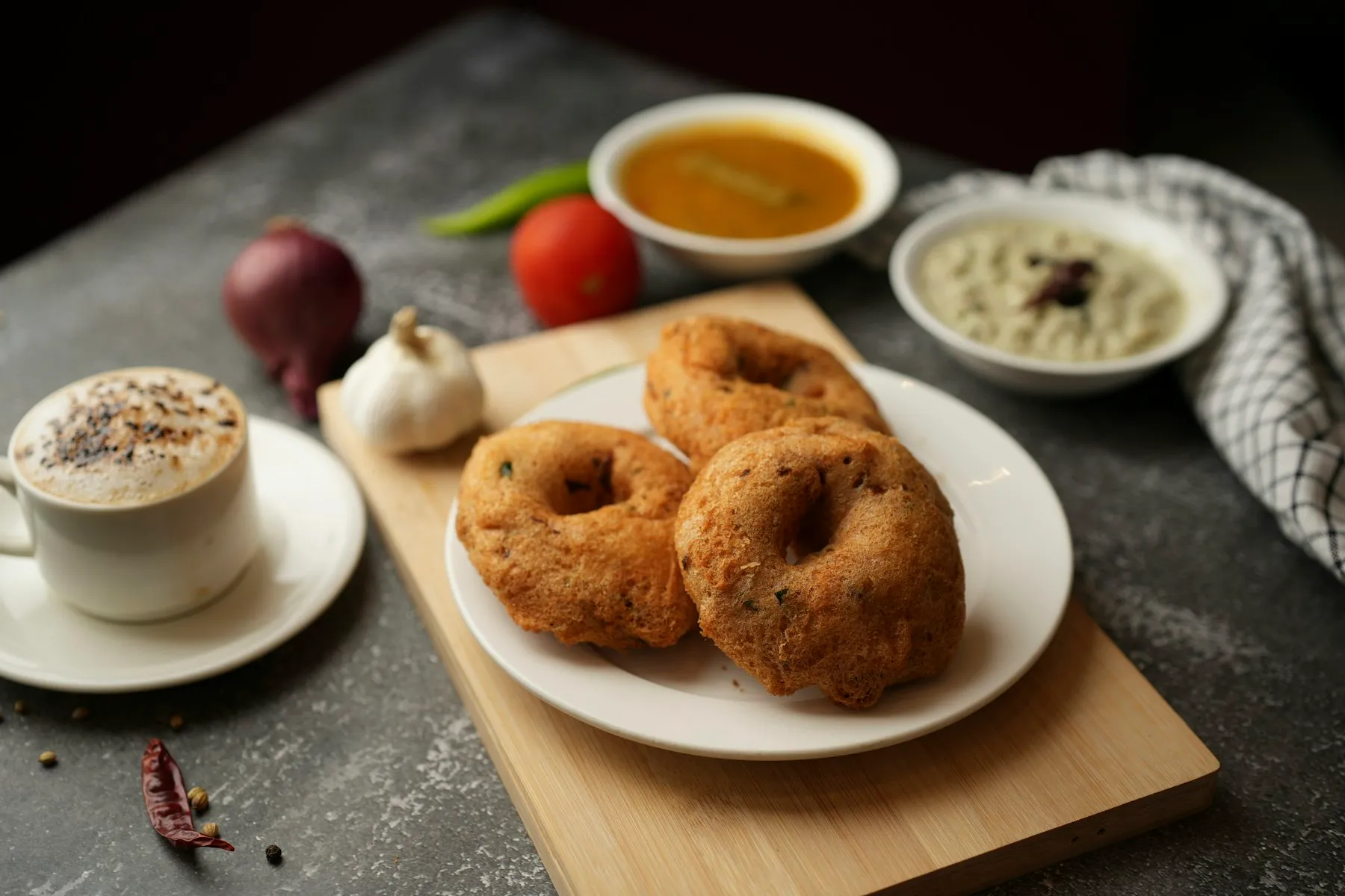 Three medu vadas on a white plate with bowls of sambar and coconut chutney and a cup of coffee