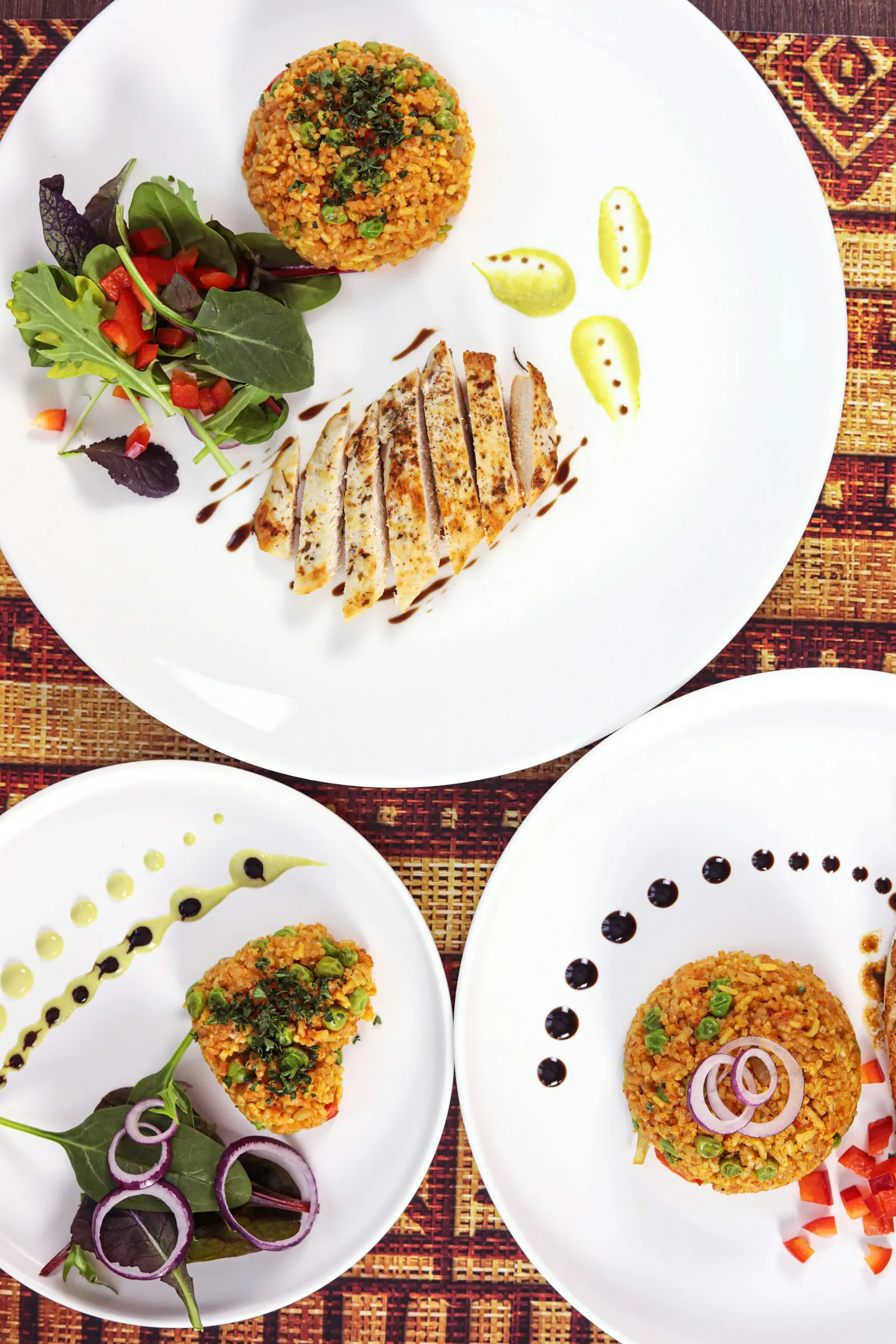 Three plates of jollof rice with grilled chicken and salad on a patterned tablecloth