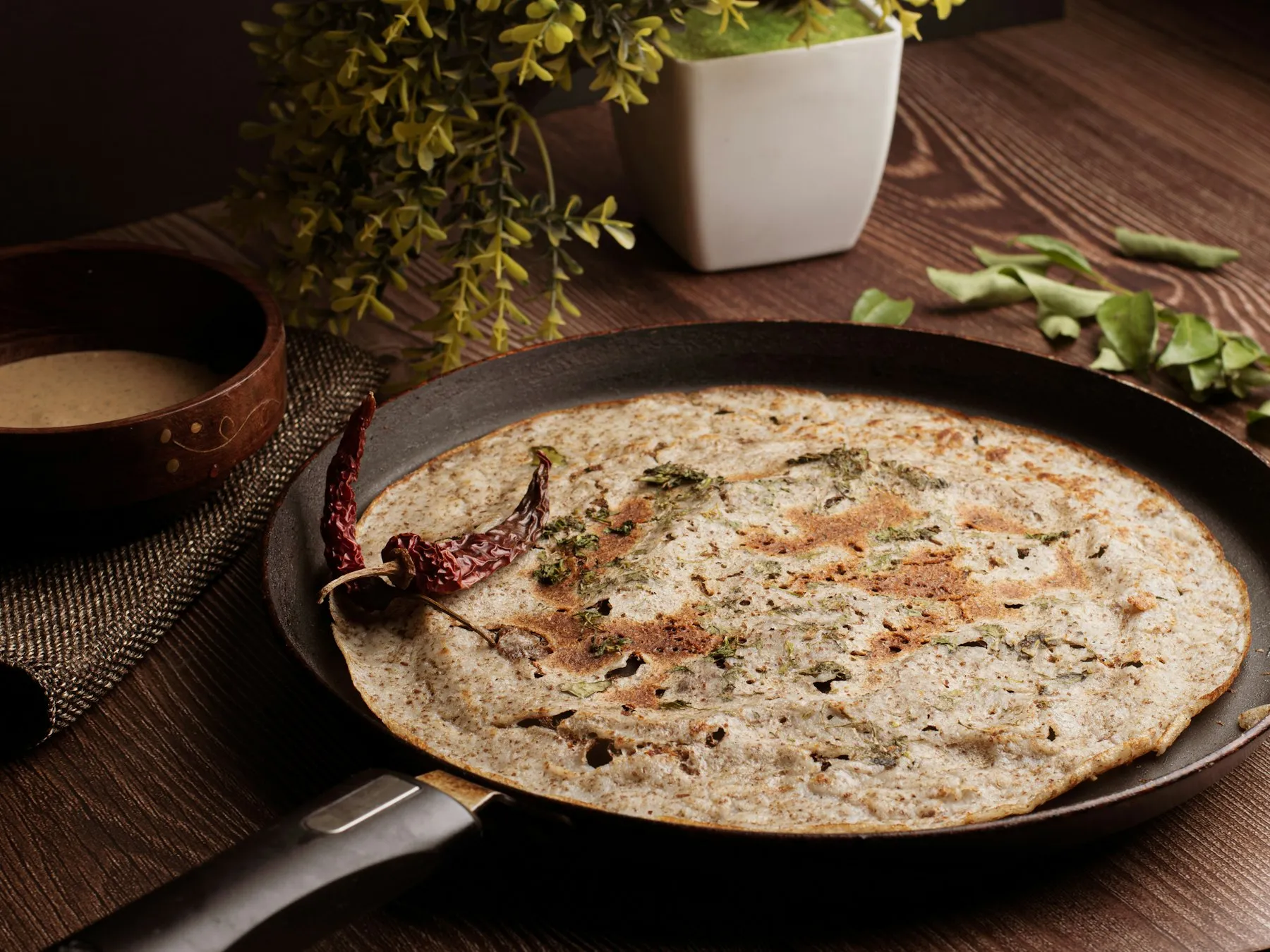 Thin savory crepe cooking in a dark pan, topped with herbs and a dried red chili