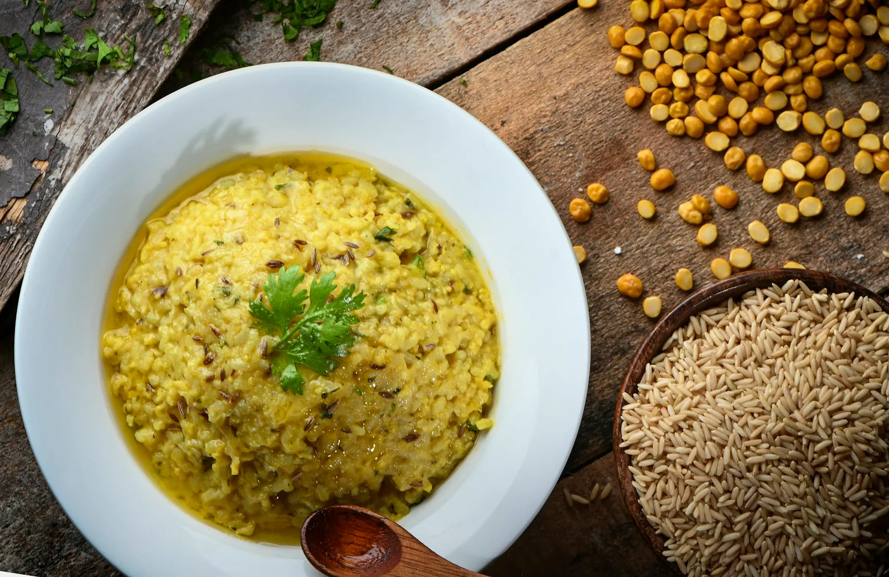White bowl of soft yellow rice and lentil porridge with cumin and a cilantro sprig, beside dry lentils and rice