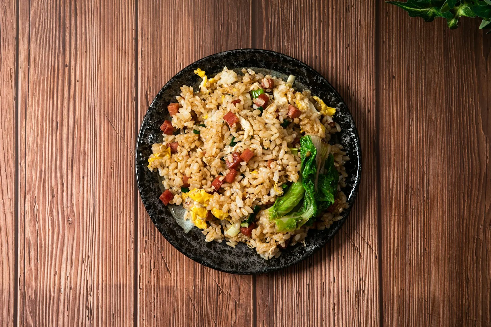 Overhead view of a dark plate of fried rice with minced ham, egg and greens on a wooden table