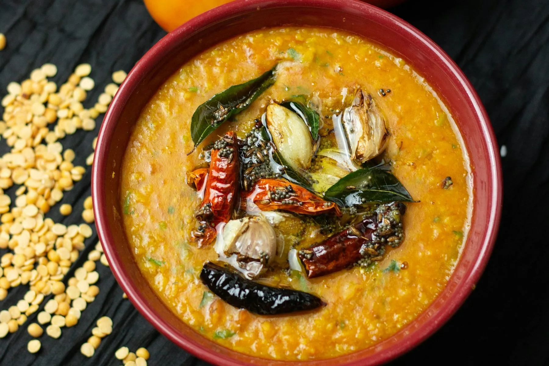 Red bowl of tomato lentil soup with a tempering of dried chilies, garlic and curry leaves beside split lentils