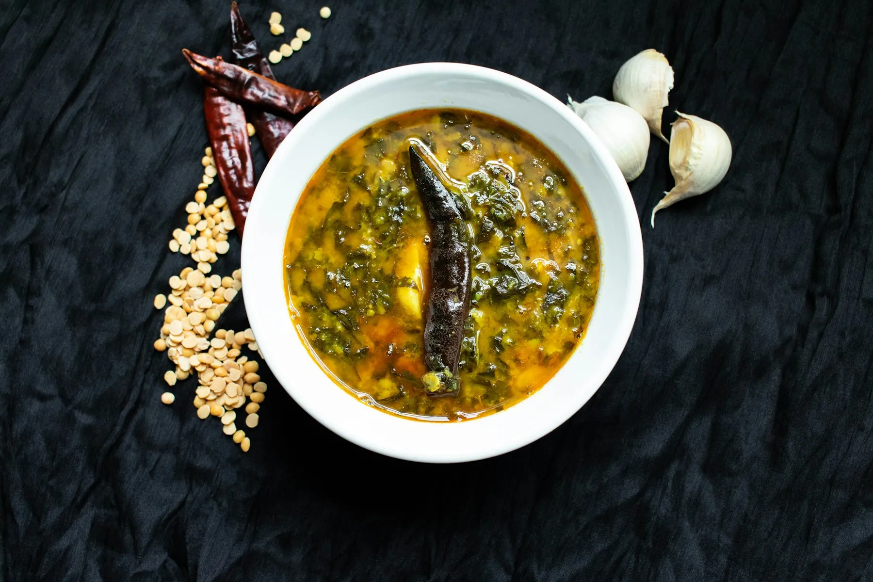 White bowl of lentil stew with greens and a drumstick, beside dried red chilies, split lentils and garlic