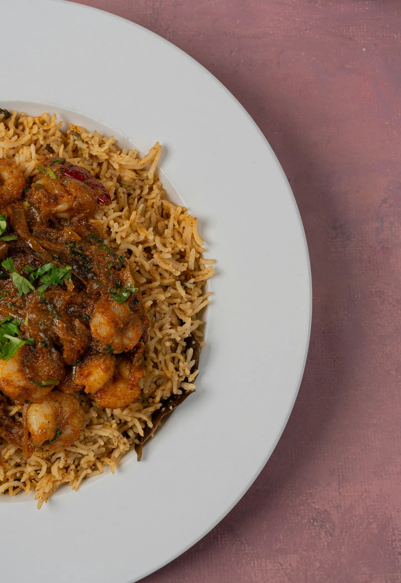 Wide white plate of spiced rice topped with prawns in a thick masala and fresh coriander