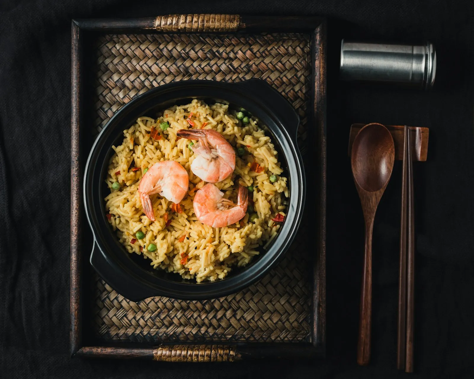 Dark bowl of fried rice topped with shrimp, with chopsticks and a spoon on a woven mat