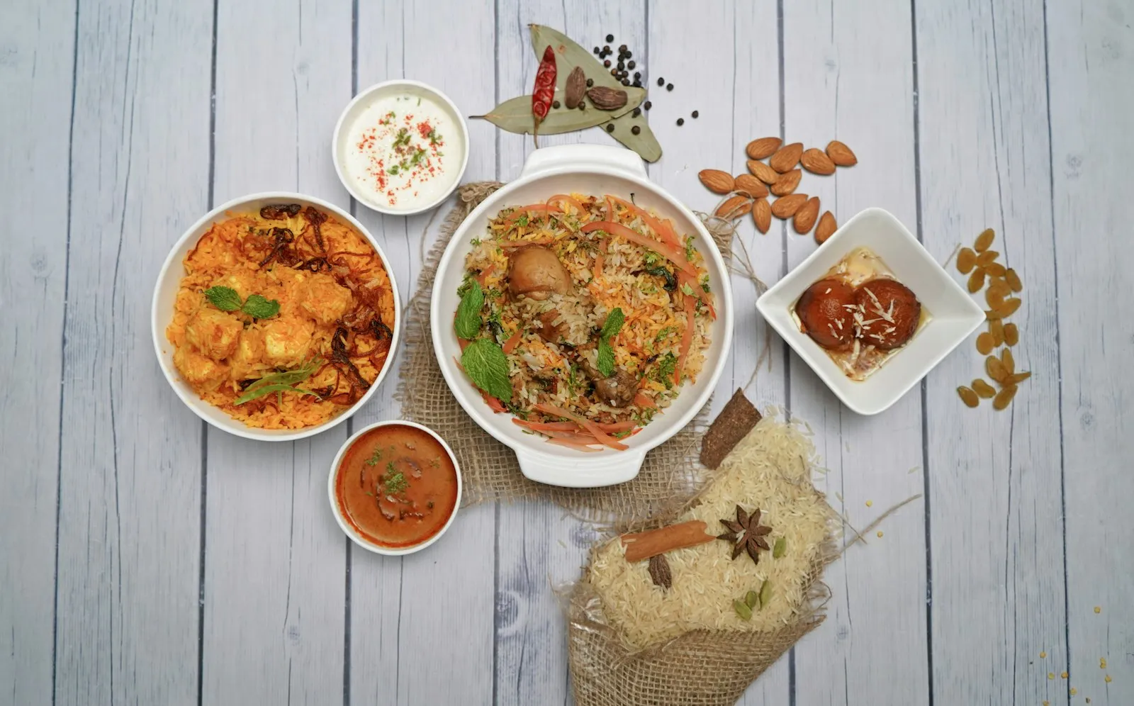 Overhead of a rice dish surrounded by small bowls of raita, curry and chutney on a white wooden table