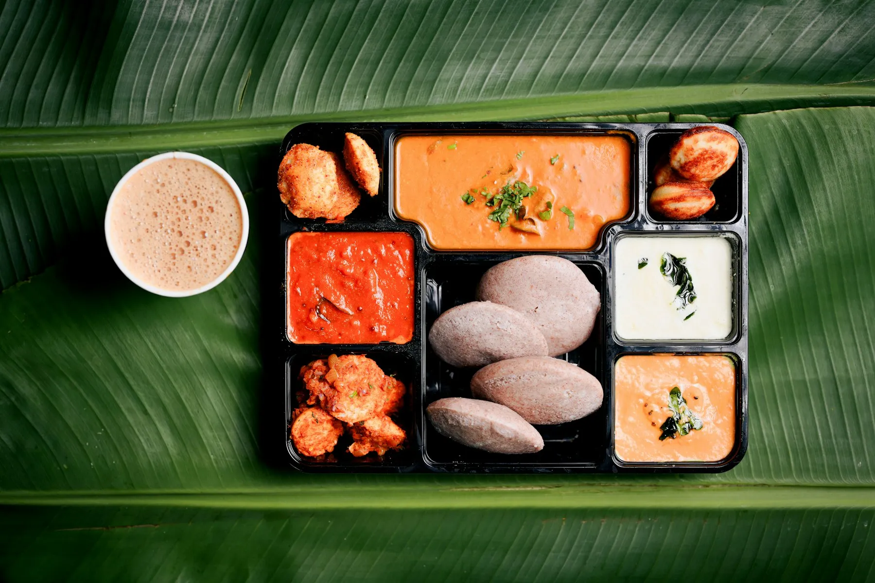 Tray on a banana leaf with idlis, fritters, sambar and chutneys, beside a cup of frothy coffee