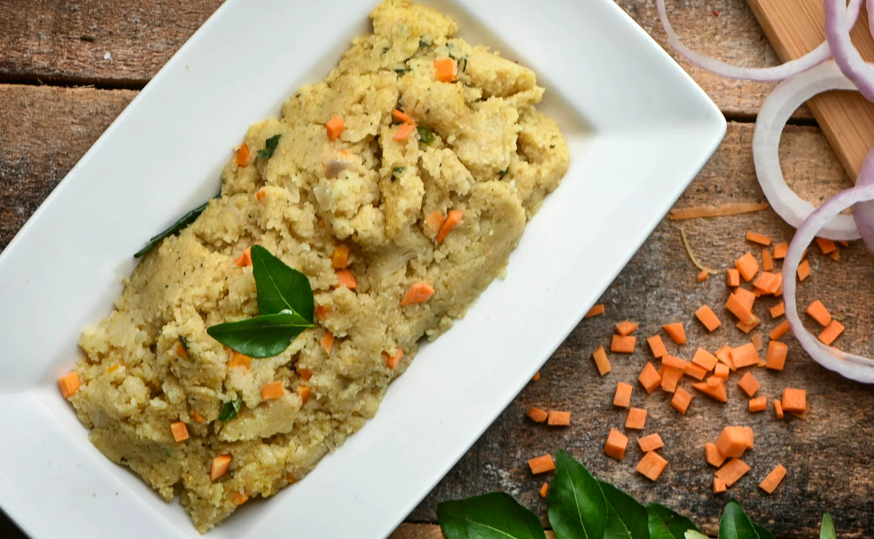 White rectangular plate of semolina upma with diced carrots and curry leaves, with onion rings and chopped carrot beside it