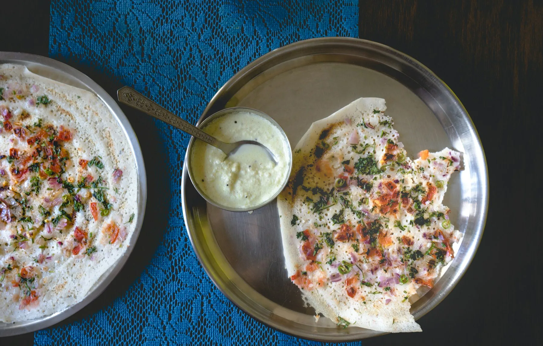 Two onion and tomato uttapams on steel plates with a bowl of coconut chutney on a blue cloth