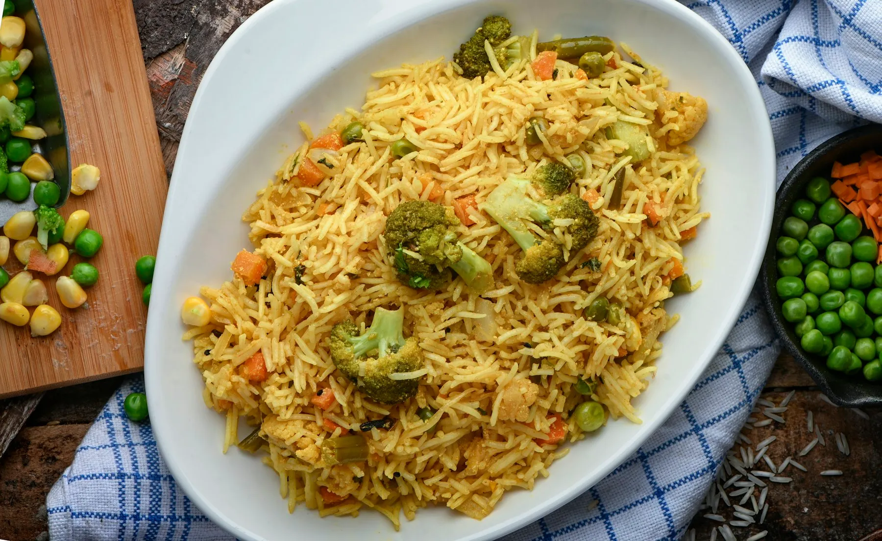 Oval white dish of spiced basmati rice with broccoli, peas, carrots and corn, with bowls of peas and corn alongside