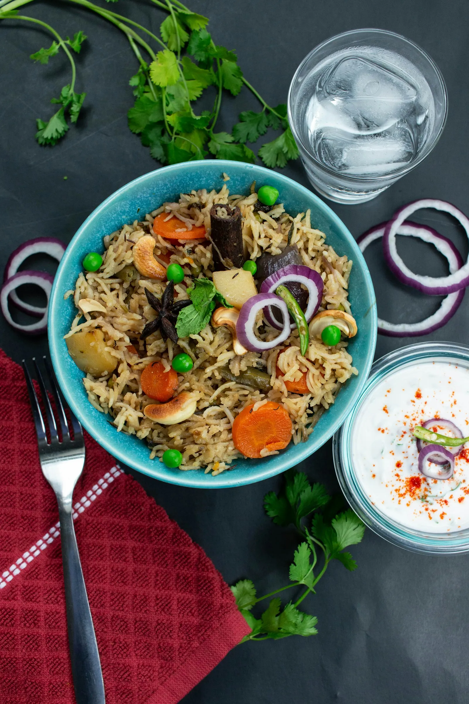 Blue bowl of vegetable biryani with peas and fried onions, served with onion rings and a bowl of yogurt